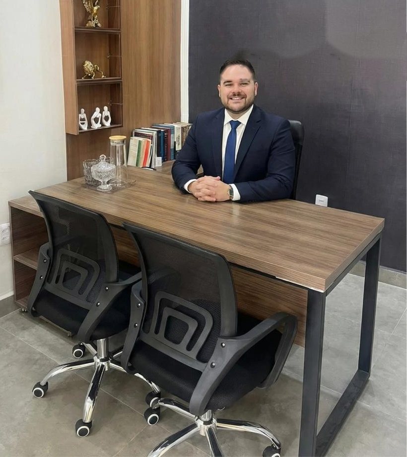 Gabriel Toledo Rezende, de terno azul-marinho, sentado à mesa de madeira do seu escritório, com estante e livros ao lado